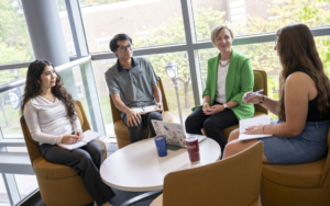 college students and a professional sit in a circle and have a discussion. they have notebooks and a laptop in front of them.