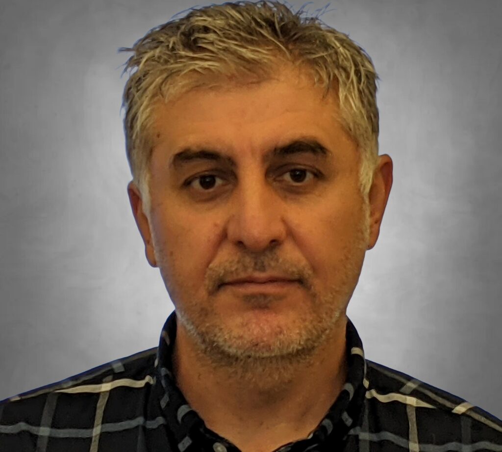 A professional headshot of a man with short hair and light facial stubble. He is wearing a dark plaid or checkered button-up shirt and is photographed against a neutral gray background with a serious, professional expression.
