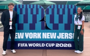 two FDU college students pose with a sign for the FIFA World Cup 2026. they are dressed in professional attire.