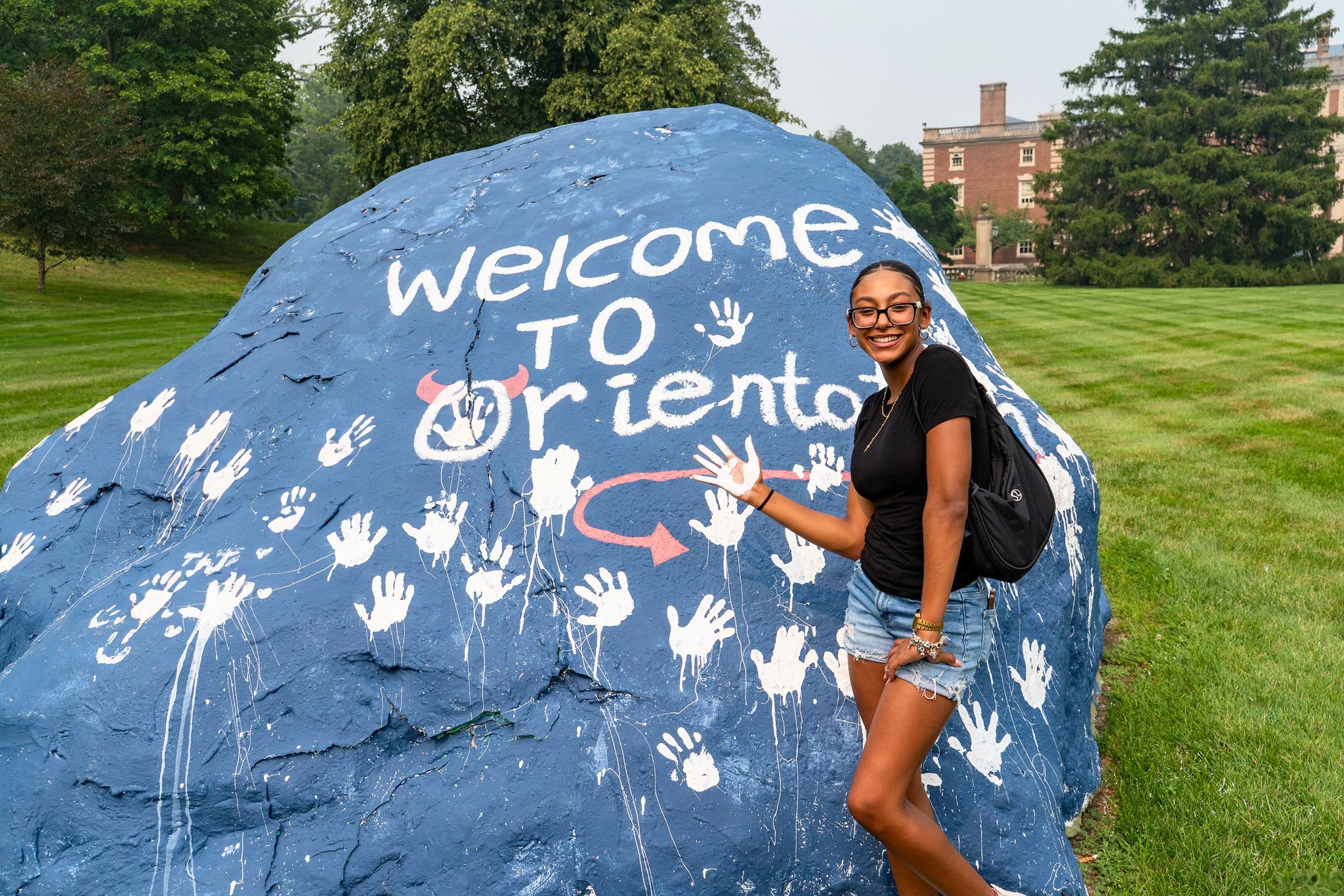 a college student leaves her handprint on a rock. the rock reads "welcome to orientation." the student smiles at the camera.