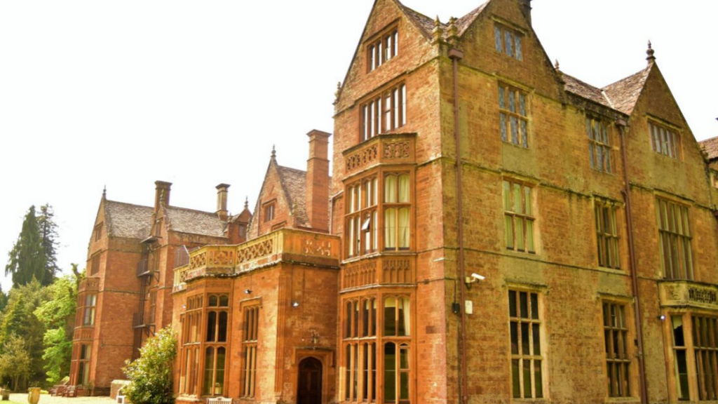 A historic red-brick manor house featuring classic English Tudor or Jacobean architecture. The multi-story building displays ornate stonework, decorative mullioned windows with leaded glass, multiple gables with steep slate roofs, carved stone balconies with intricate balustrades, and tall brick chimneys. The warm-toned brickwork shows characteristic patterns and decorative details. Mature trees and landscaping surround the building, which appears to be set in park-like grounds under a bright sky.