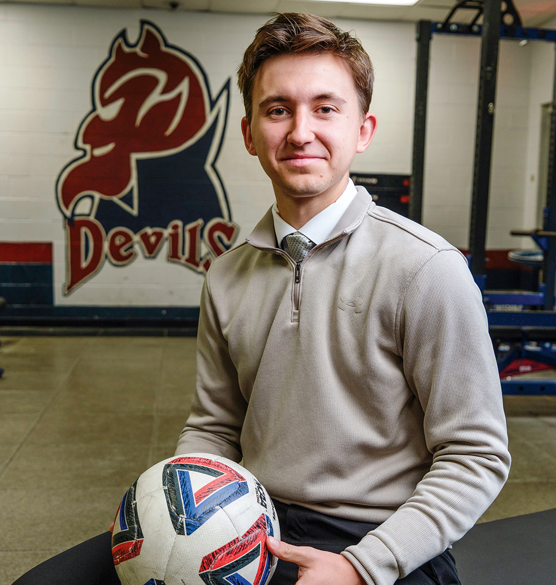 A young man sits in a gym weight room, holding a soccer ball.
