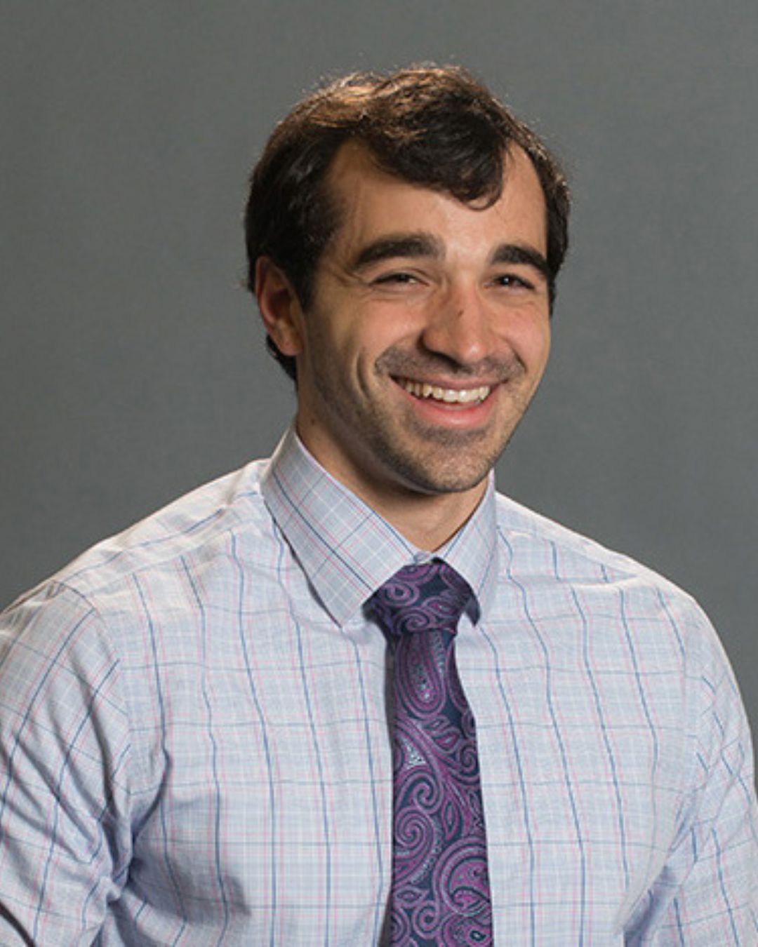 a headshot of a man. he wears a button-down shirt with a purple tie.