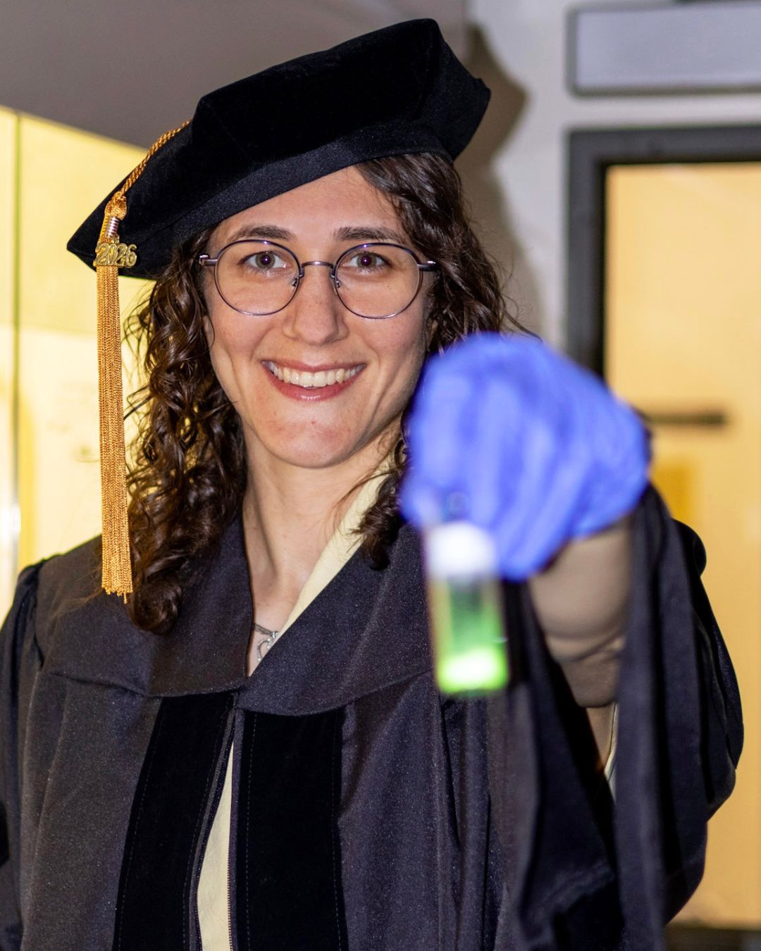 a headshot of a woman. she wears academic regalia and holds up a vial.