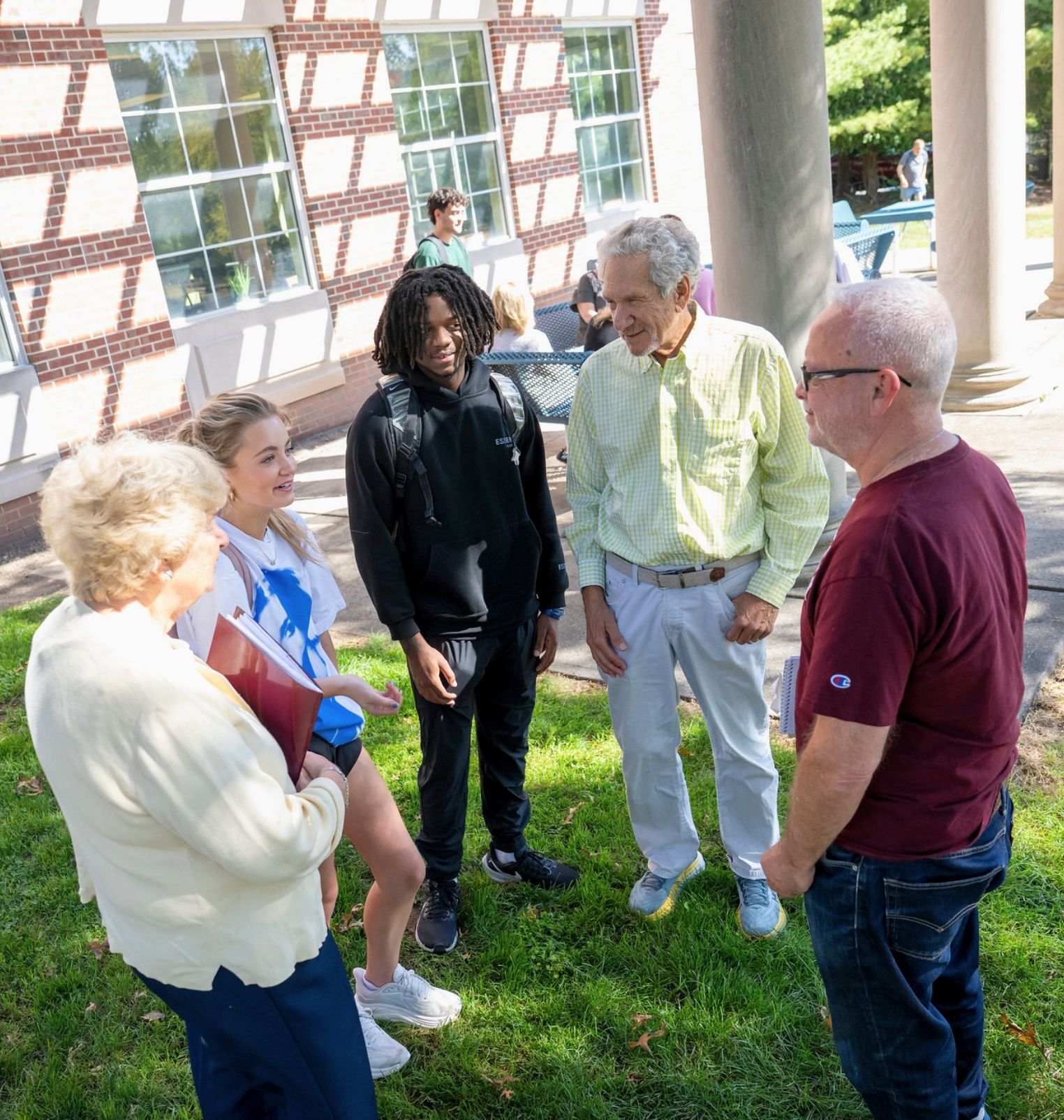 a group of college students stand in a circle outside and talk amongst themselves. some of the students are traditional college age, while some are more elderly.