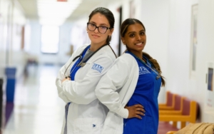 two students stand back to back. they both wear nurse uniforms and lab coats. one student crosses her arms and the other student has her hands on her hips.
