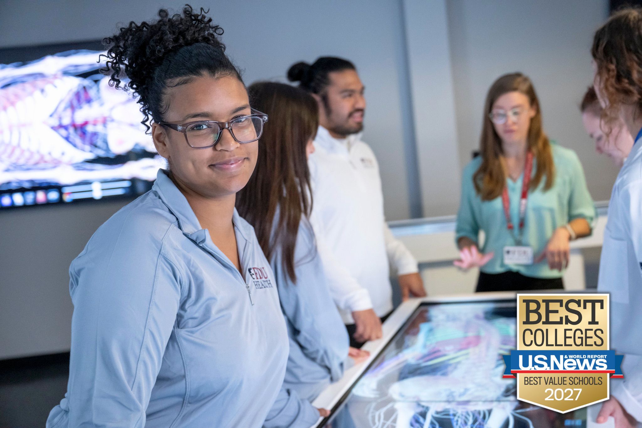 a college student wears a quarter-zip jacket with the FDU Health logo. she smiles at the camera. behind her, students and a professor work on a screen with a digital model of a human skeleton muscle system.
