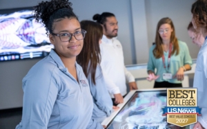 a college student wears a quarter-zip jacket with the FDU Health logo. she smiles at the camera. behind her, students and a professor work on a screen with a digital model of a human skeleton muscle system.