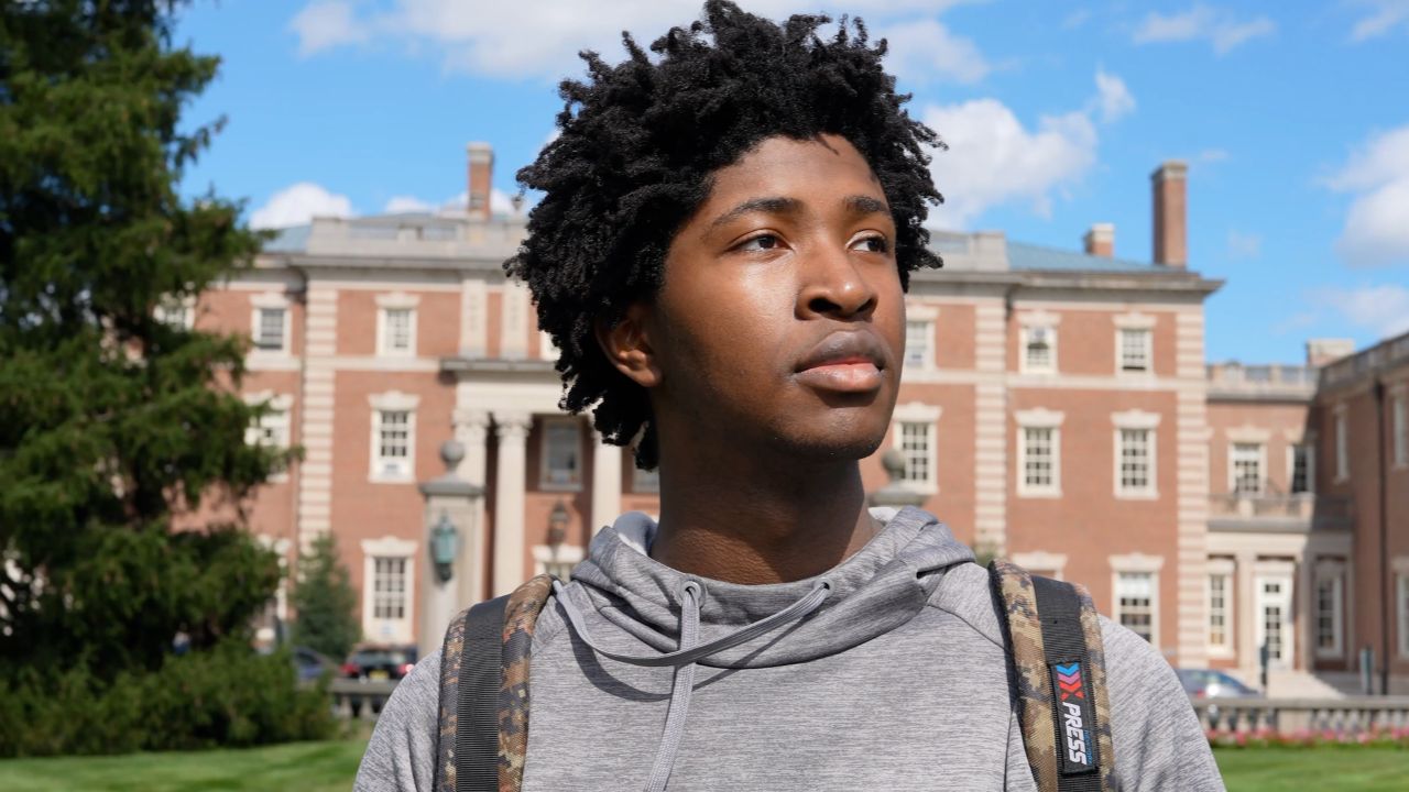 a close-up of a student looking off to the distance. he wears a hoodie and a backpack. the FDU Hennessy Hall Mansion stands behind him.