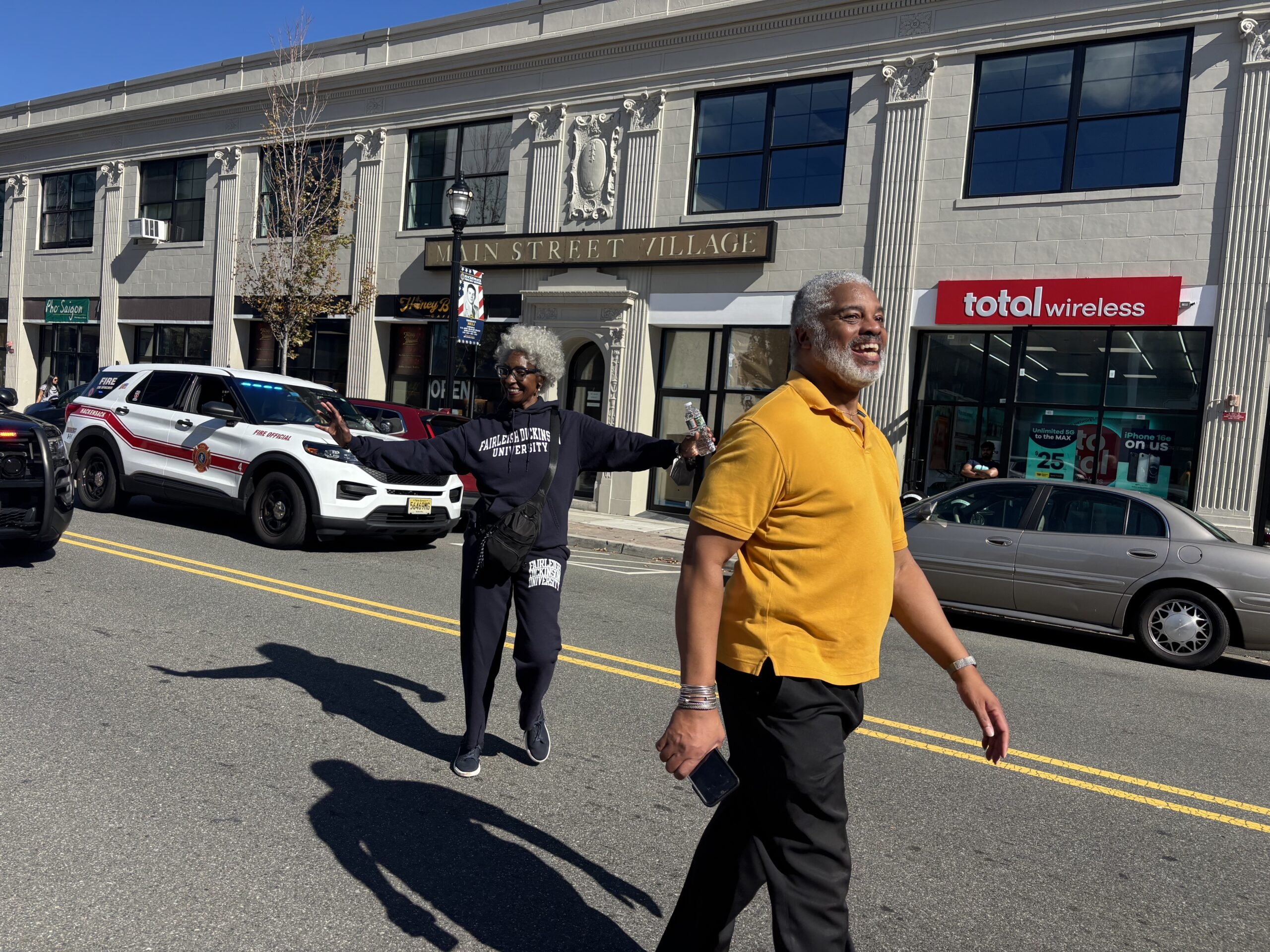 A man in a yellow shirt and a woman in a Fairleigh Dickinson University sweatshirt walking down a street with a fire official vehicle and Main Street Village storefronts in the background.